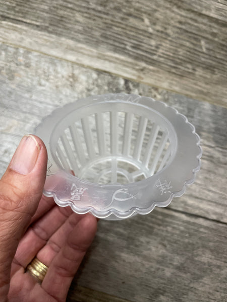 A hand holding a 4” scalloped edge clear plastic orchid basket.. top view