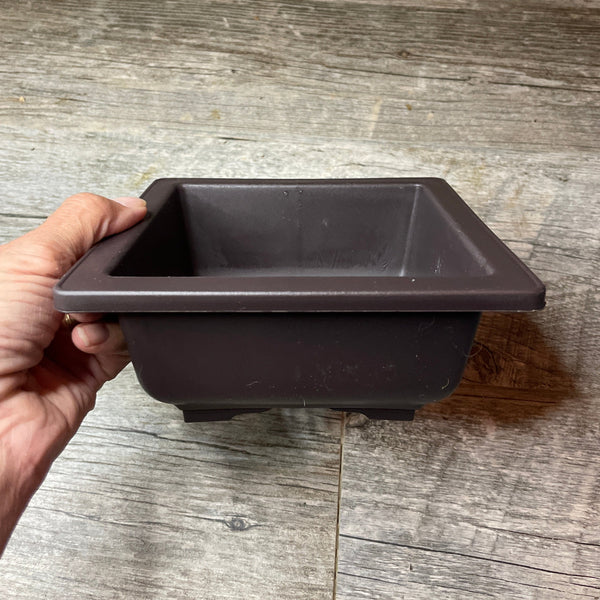 a hand holding a square 6.6" brown plastic bonsai pot - side view