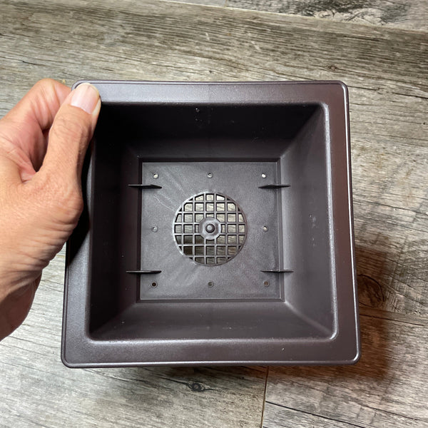 a hand holding a brown plastic bonsai pot with a view into the bottom of the pot showing the drainage holes.